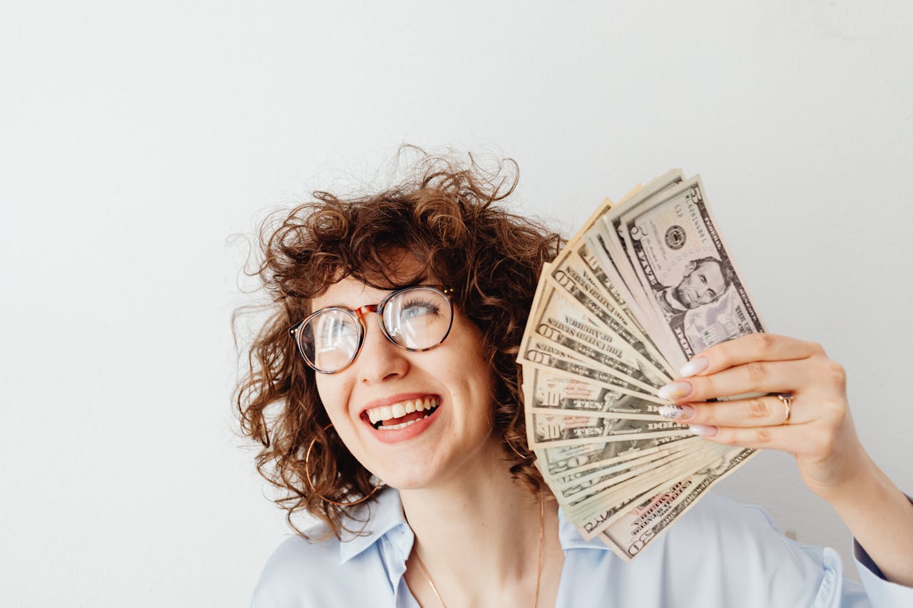 Happy woman with curly hair and glasses holding US dollar bills against a white background.